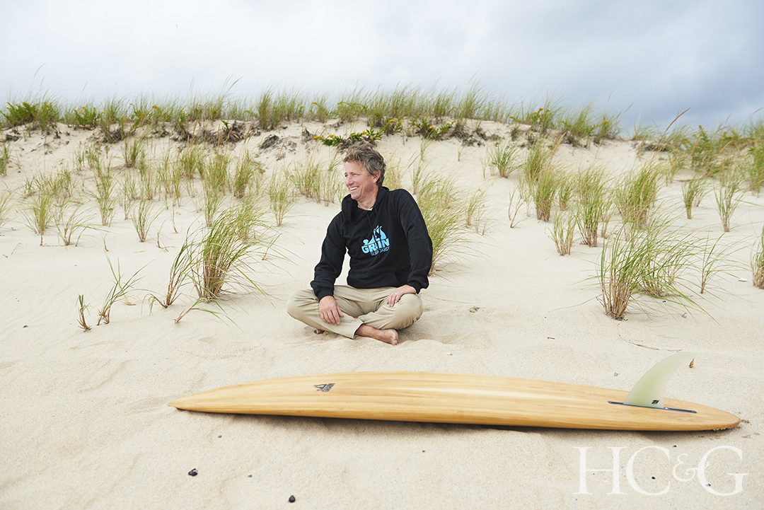 Man sitting cross legged in the sand in front of wooden surfboard.