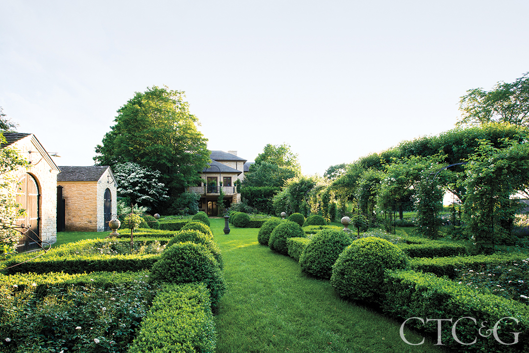 A vibrant green garden is flanked by a stone home and garden sheds.