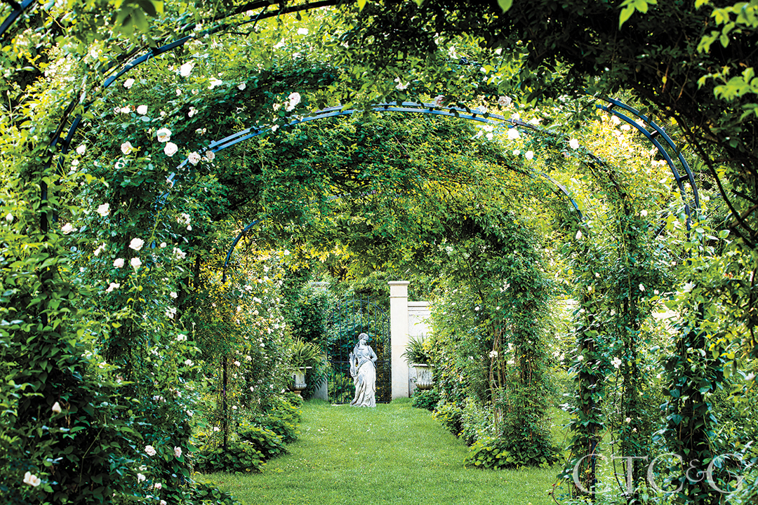 High archways are covered with climbing white roses, framing a white statue of a woman.