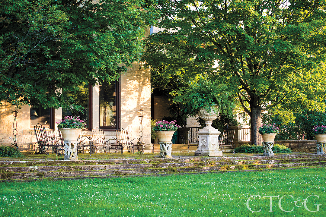A sunny stone terrace with chairs and potted flowers, shaded by trees.