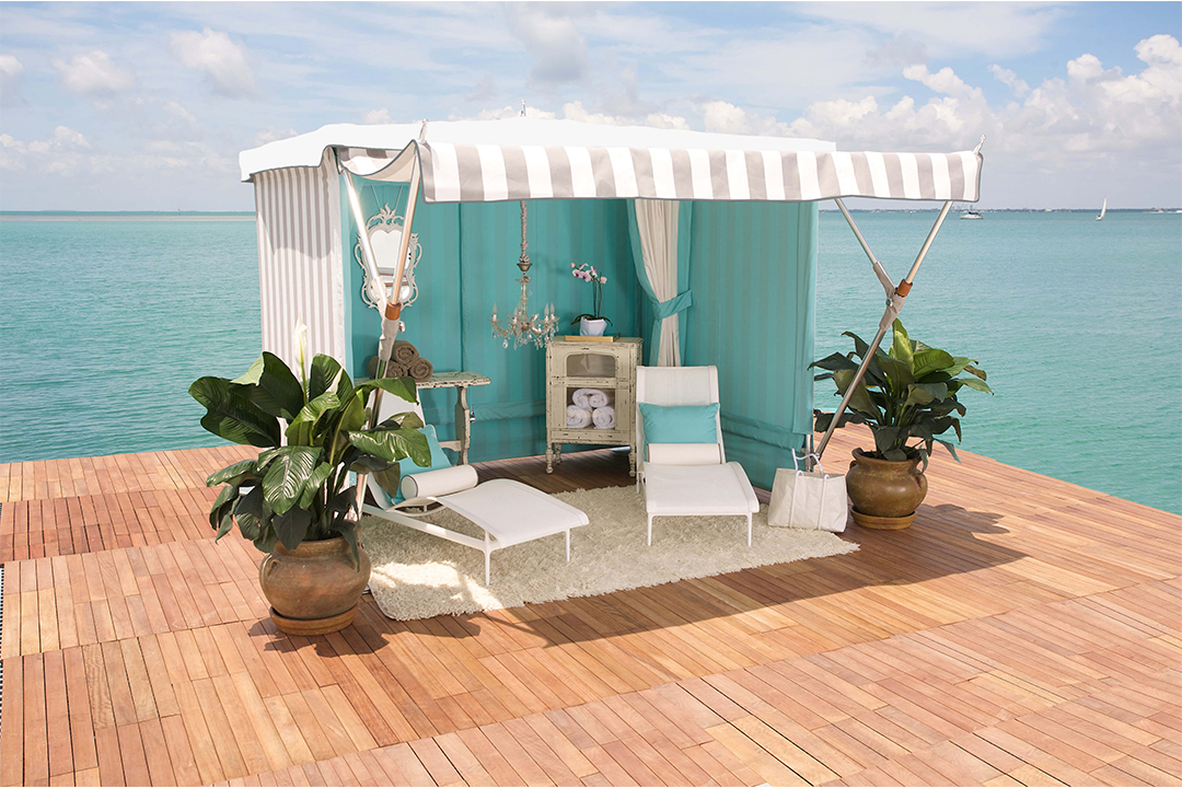 A wooden dock with a seaside pavilion, featuring a white striped canopy, white lounge chairs, a white carpet, and large potted plants.
