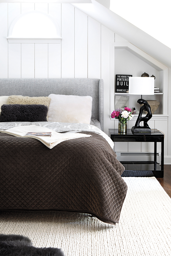 linen and wood upholstered bed in primary bedroom; table lamp and floral arrangement