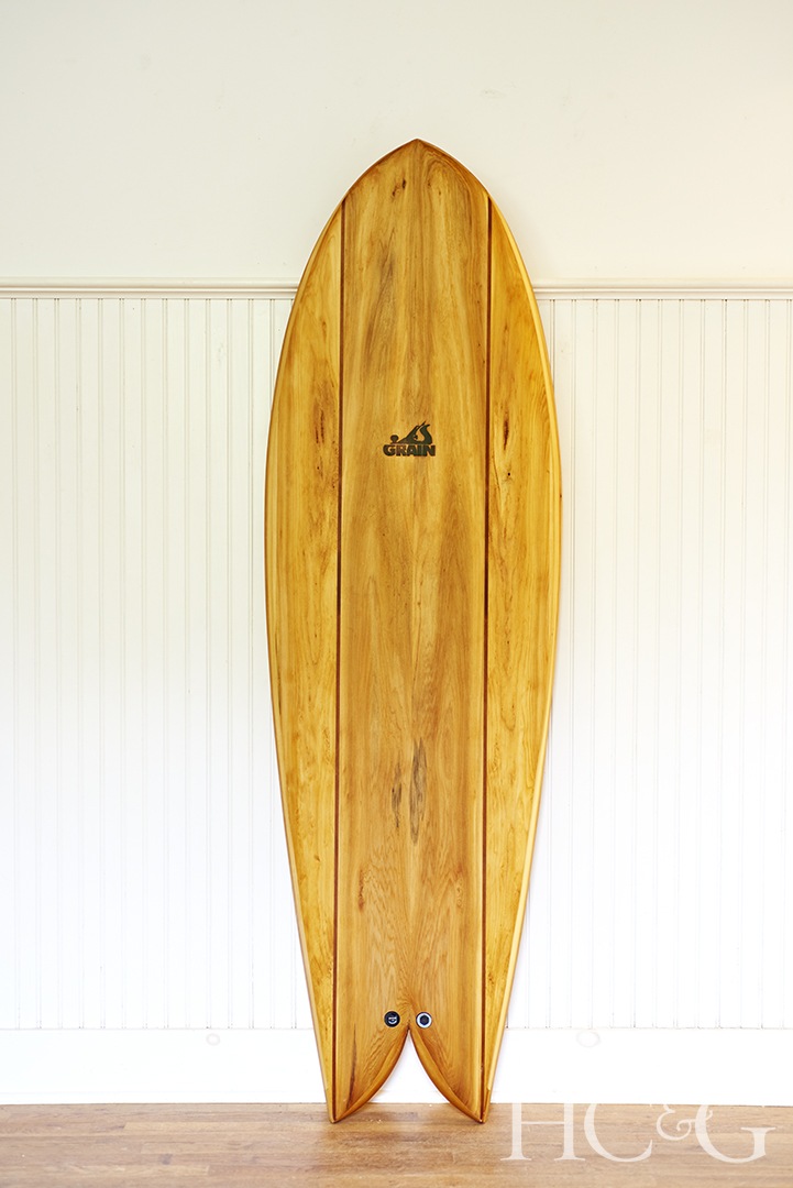 A finished wooden surfboard proper up vertically against a white wall.
