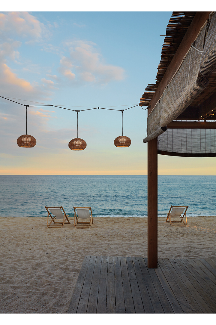 A garland of three woven light fixtures hangs in front of a beach at sunset.