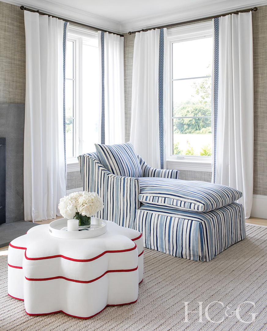 A blue and white striped lounge sits near a bright large window. A white ottoman with red lining is to the left.
