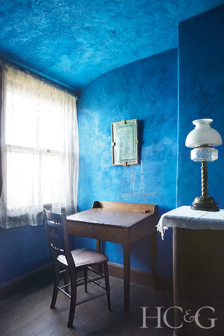 A rich blue room with a bright window on the left featuring a wooden desk and chair.