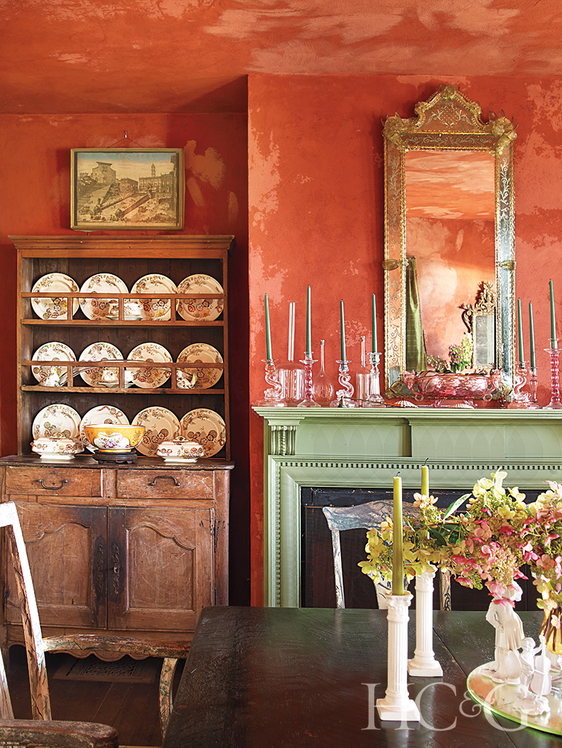 An orange wall with a wooden plate shelf backs a wooden dining table. A green fireplace frame is to the right of the shelf.