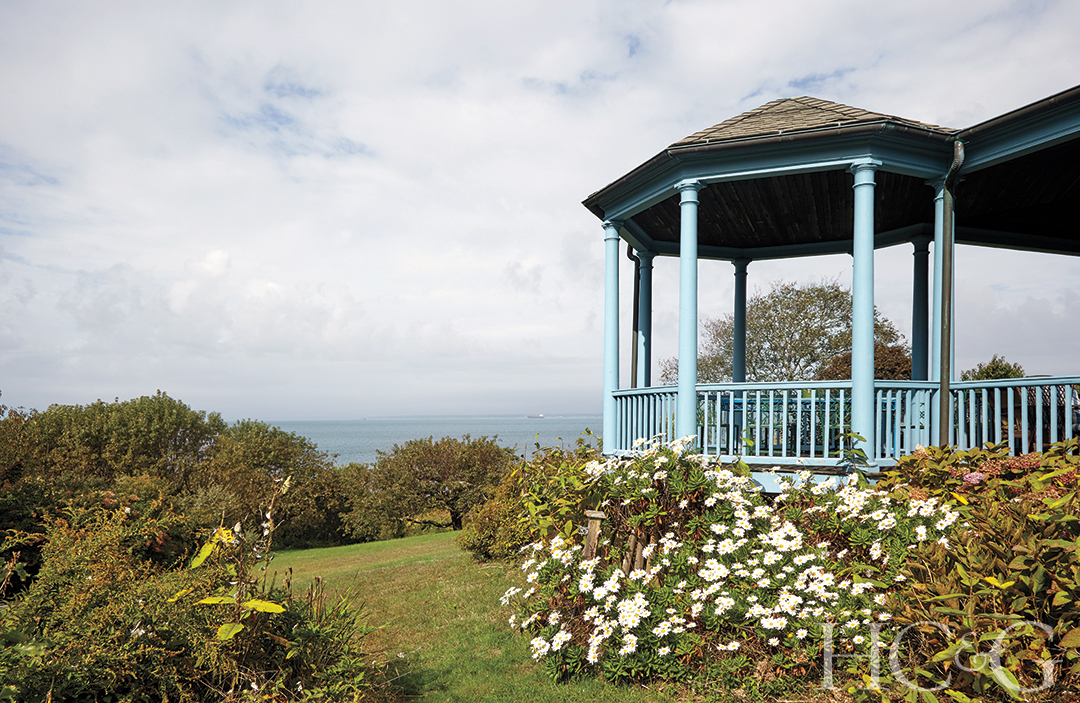 A baby blue gazebo sits with the water in the distance, surrounded by greenery.