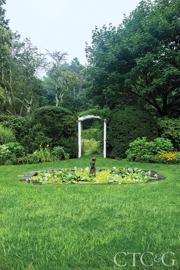 A fountain filled with green sits before a white arch leading into more greenery.