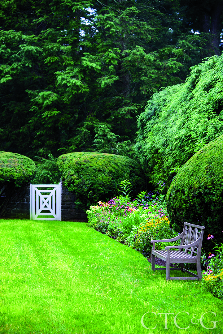 A white fence leads into a garden with a wooden bench and flowers.