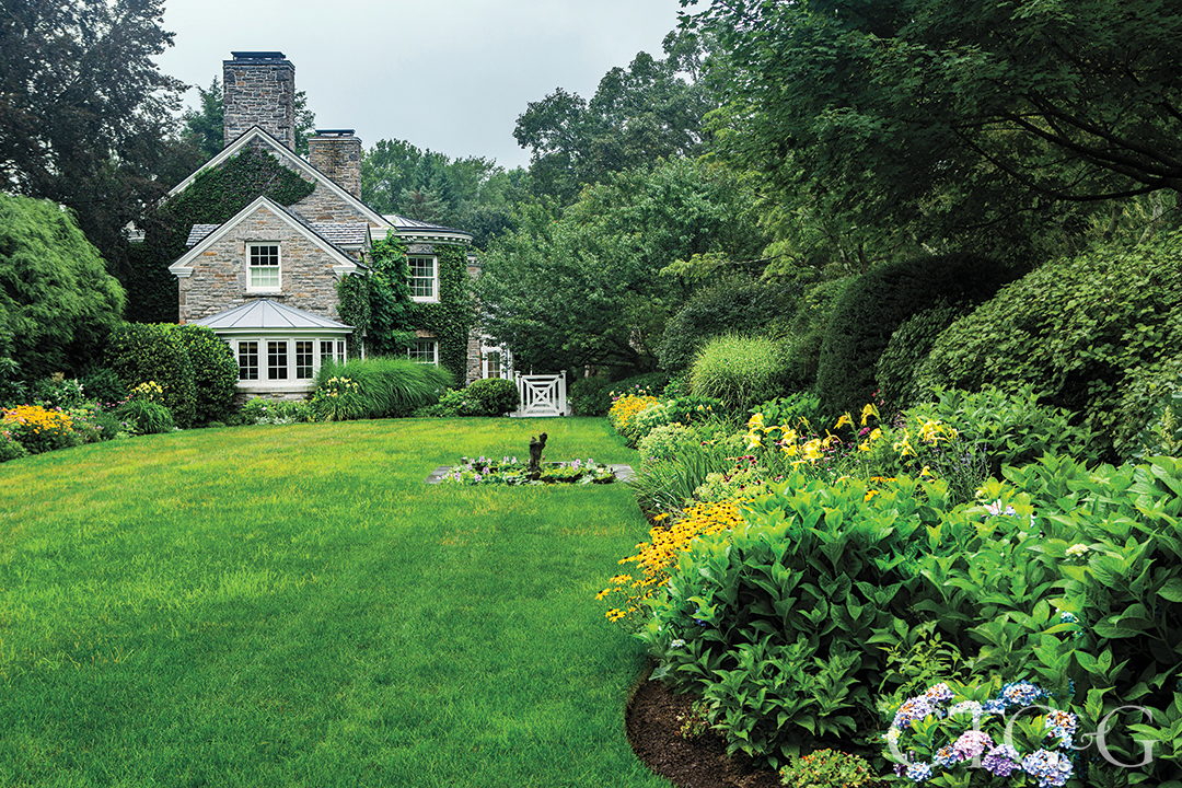 A stone house partially covered in greenery sits behind a green lawn and garden.