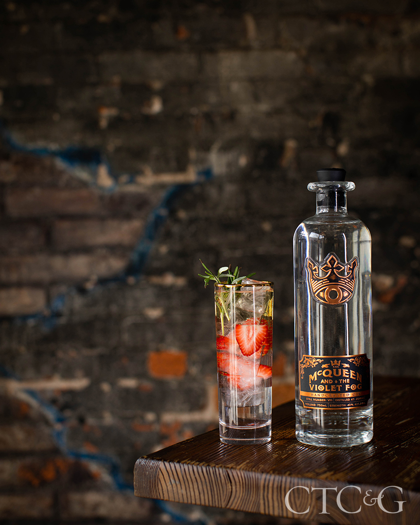 A clear bottle of gin sits to the right of a small drink with strawberries and greens.