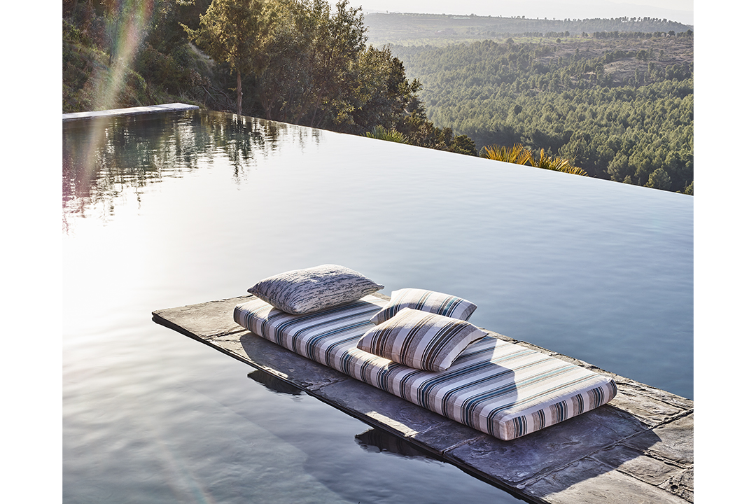 A long striped cushion and three pillows sit on a stone runway, surrounded by an infinity pool.