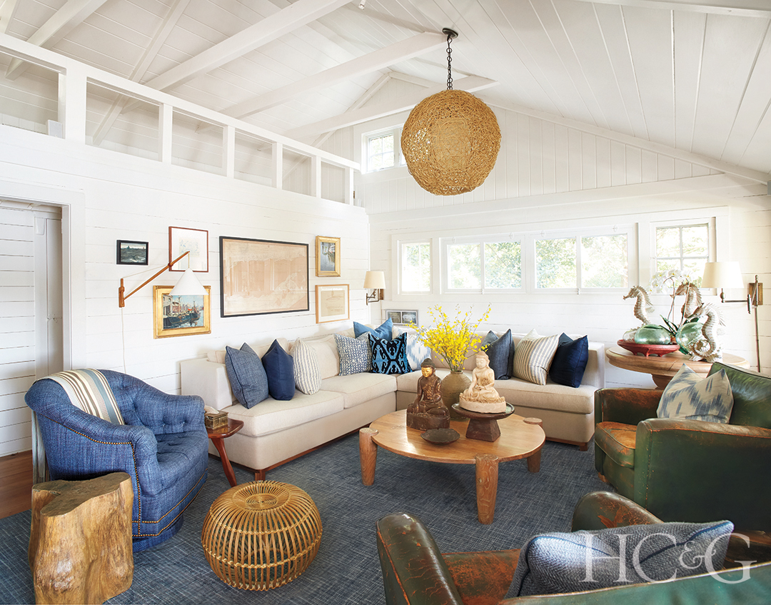 A circular lighting fixture hangs over a white walled living room with a blue carpet. A sofa studded with blue pillows sits behind a wooden table.