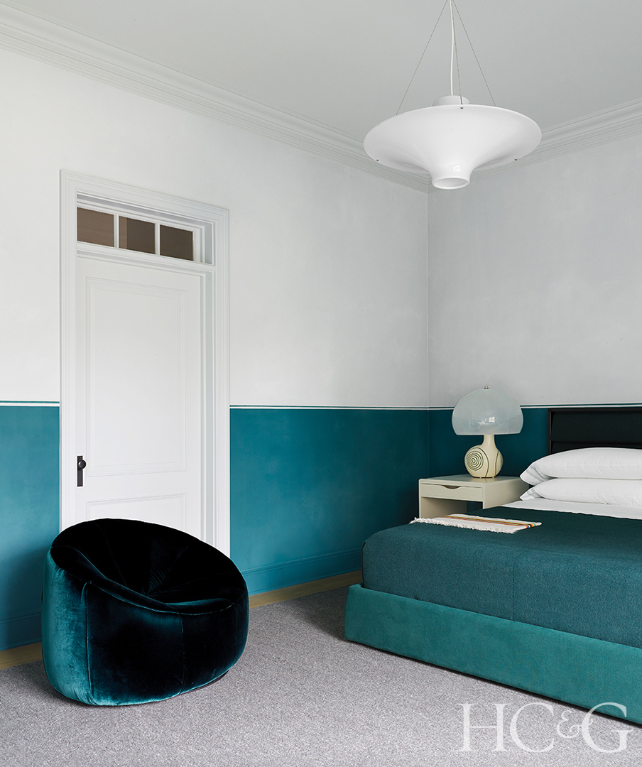 A teal velvet lounge chair sits in the corner of a white and teal bedroom.