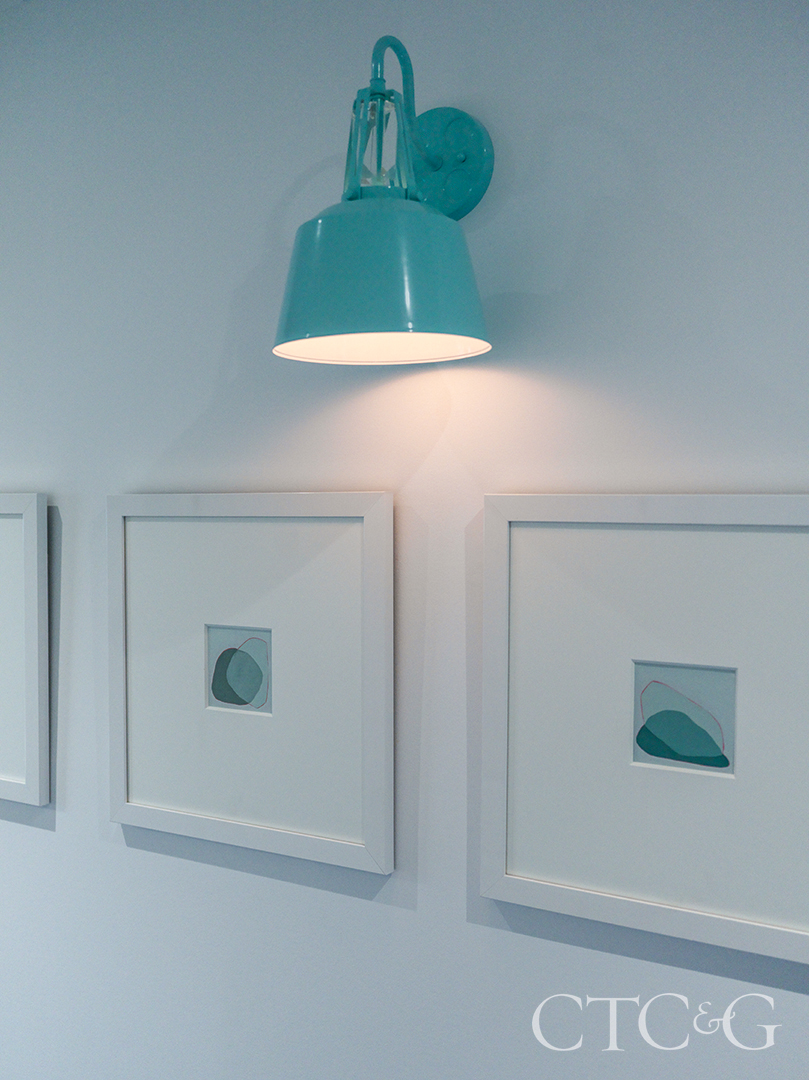 trio of Lisa Carroll paintings hang below blue sconce in hallway